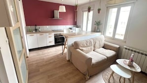 Une cuisine moderne avec des meubles blancs, un plan de travail en bois et des plantes suspendues. On y trouve un canapé beige et une petite table ronde sur laquelle est posé un vase.