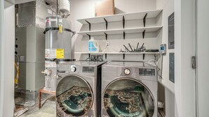 Un espace buanderie comprenant deux machines à laver à chargement frontal, un chauffe-eau et des étagères au-dessus pour le rangement.