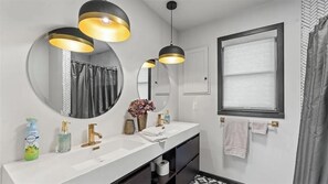 Un baño moderno con un espejo grande, dos lámparas colgantes, un lavamanos blanco y una ventana con cortina.