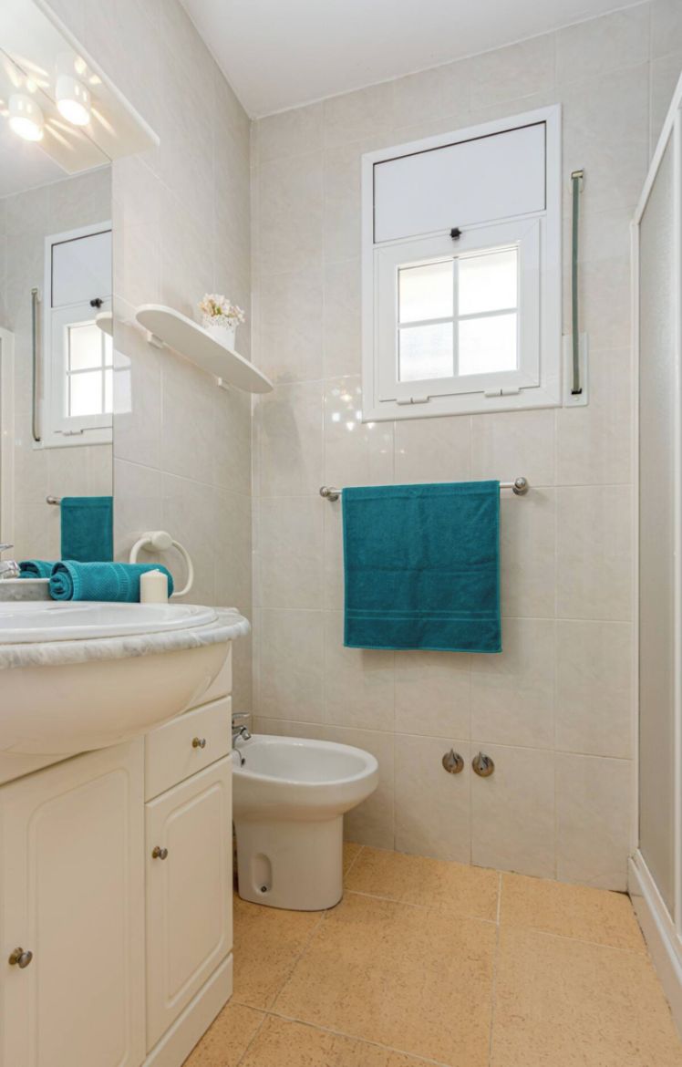 Casa de banho com toucador branco, bidé, porta-toalhas com toalha azul e janela sobre o sanitário.