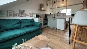 A modern kitchen with white cabinets, a microwave, and a sink. There is a green sofa and a small round table with a white furry rug. - Studio Les Tulipes (Calais)