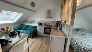 A compact kitchen with a microwave, sink, and stove. There is a small refrigerator, a teal sofa, and a dining table with a vase of flowers. - Studio Les Tulipes (Calais)