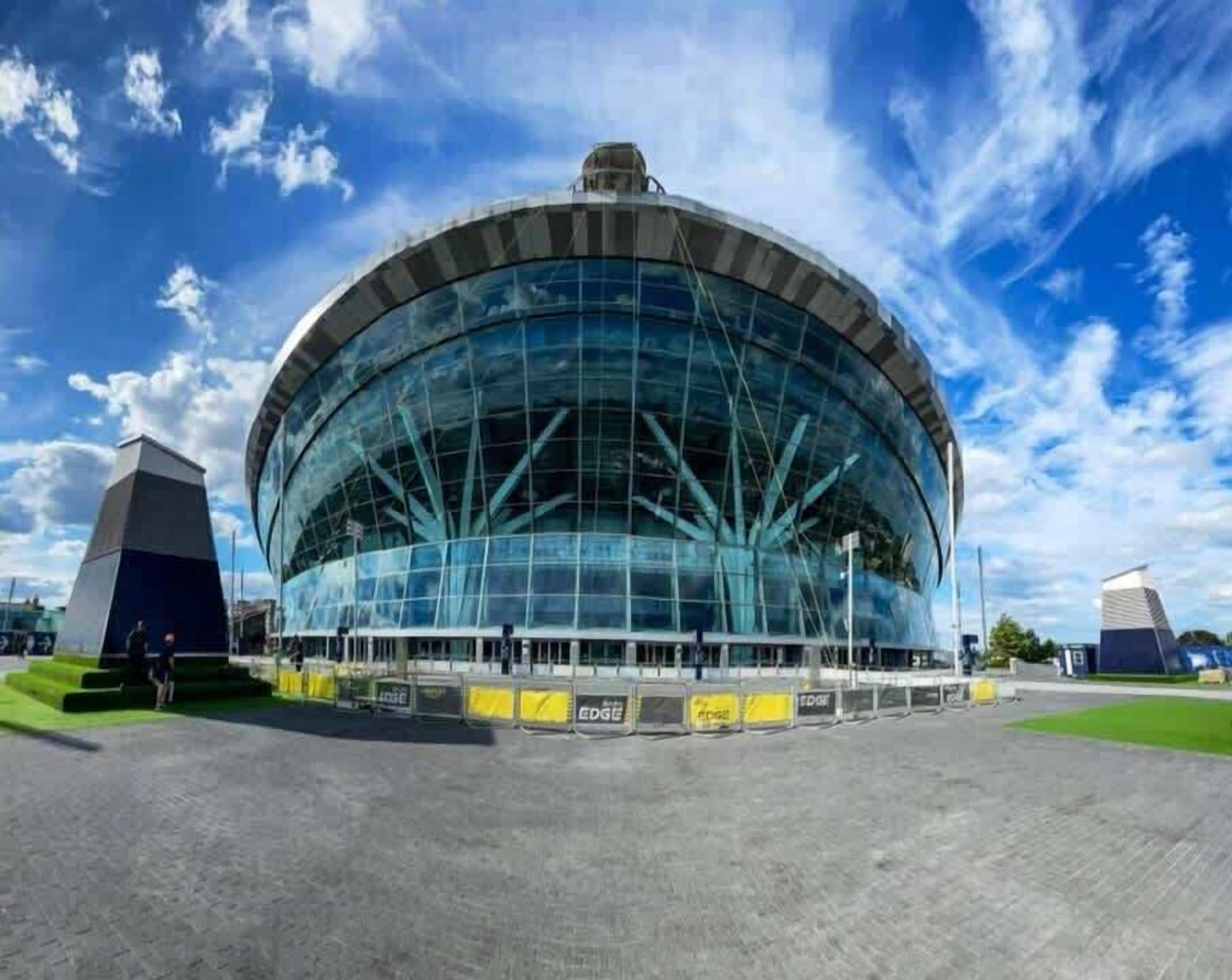A large, circular building with a glass facade and a central dome, surrounded by a paved area and a smaller structure to the left.