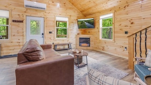 A cozy cabin interior with a leather sofa, a fireplace, a television, and large windows offering a view of the outdoors. - Riverfront Cabin~Firepit~Private Boat Ramp w/ river access~Dog-Friendly! (Jamestown)