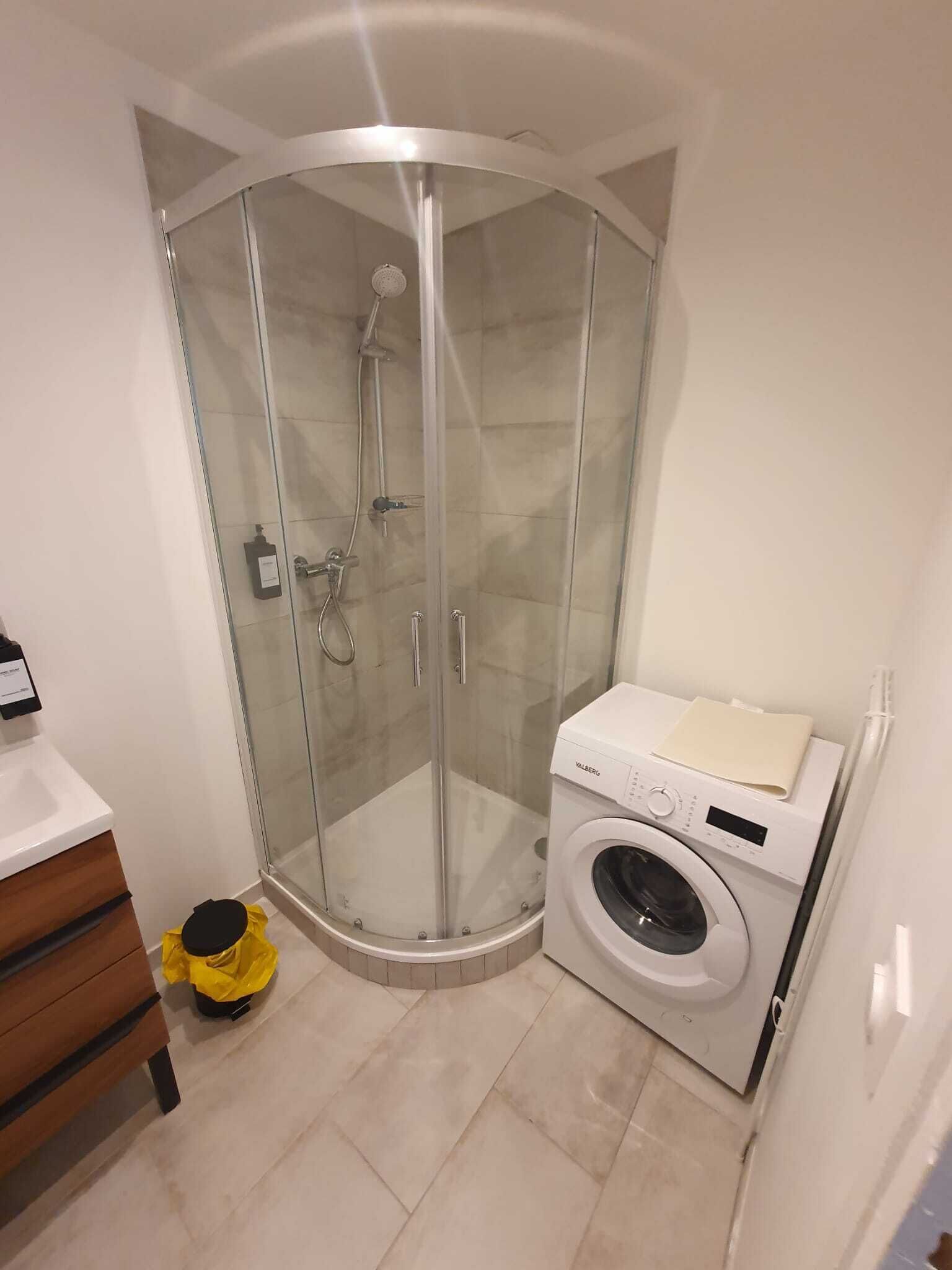 A corner bathroom with a glass shower enclosure, a washing machine, and a yellow bucket.