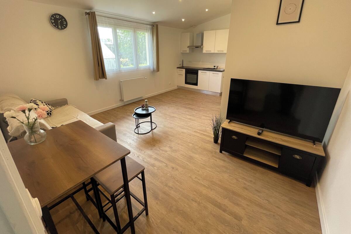 Uma sala de estar moderna com piso de madeira, televisão, mesa de jantar e cozinha ao fundo.