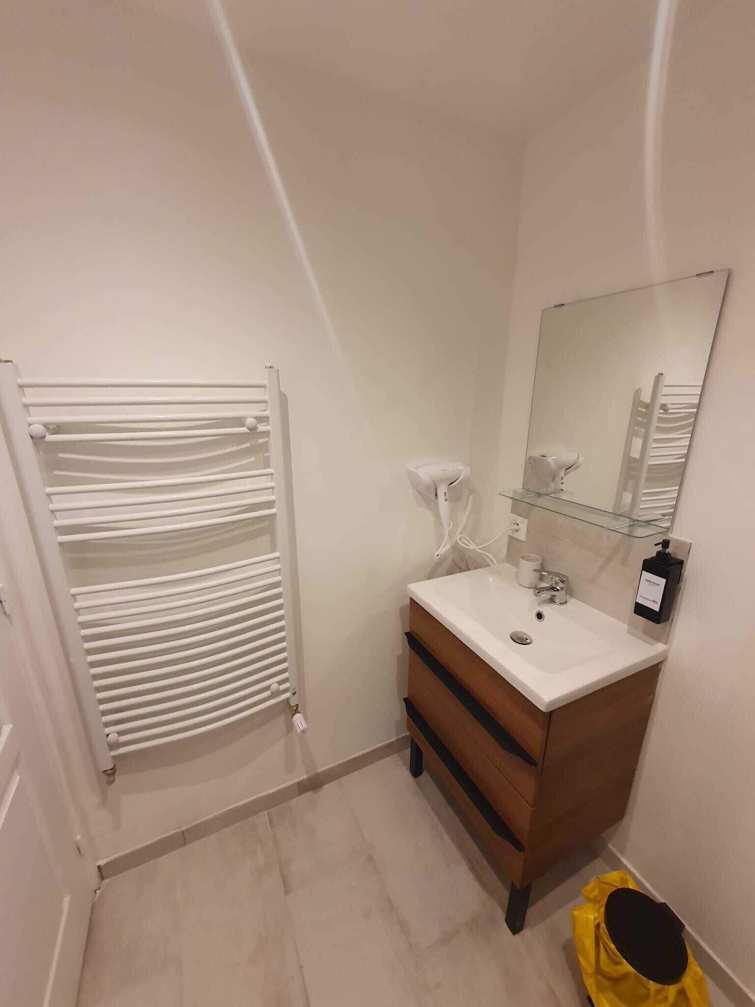 Un baño con un radiador blanco, un lavabo con un espejo encima y un bolso amarillo en el suelo.