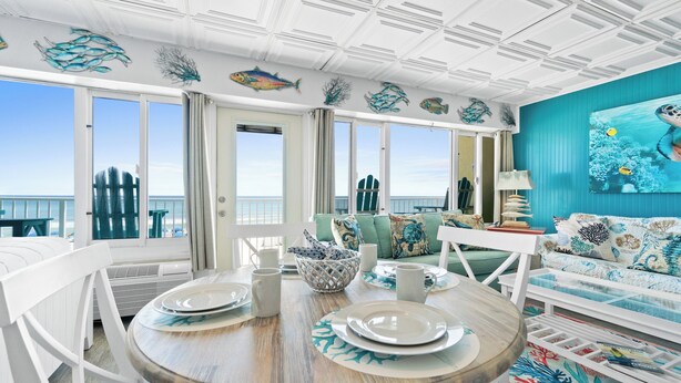 A dining area with a round table set for four, a sofa with a view, and a mural of sea life on the wall. - Continental by Book That Condo (Panama City Beach)