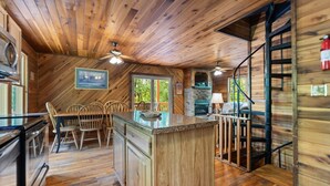 Interno di una baita in legno con angolo cottura dotato di isola centrale, tavolo da pranzo in legno, sedie e camino in pietra.