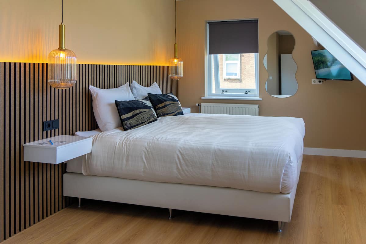 A modern bedroom with a large bed, a bedside table, a hanging light fixture, a wall-mounted TV, and a window with a view of the outside.