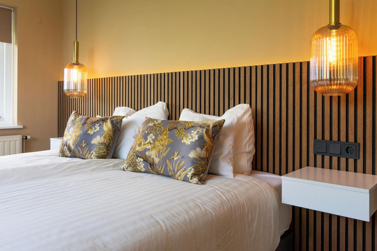 A neatly made bed with floral pillows, a bedside table, and a wall-mounted power outlet.