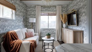 A room with a wooden ceiling, floral wallpaper, a sofa with a brown blanket, a small round table with a plant, and a TV on the wall. - Charming Cottage on the Boise Bench. Minutes from downtown, BSU & the airport. (Boise)