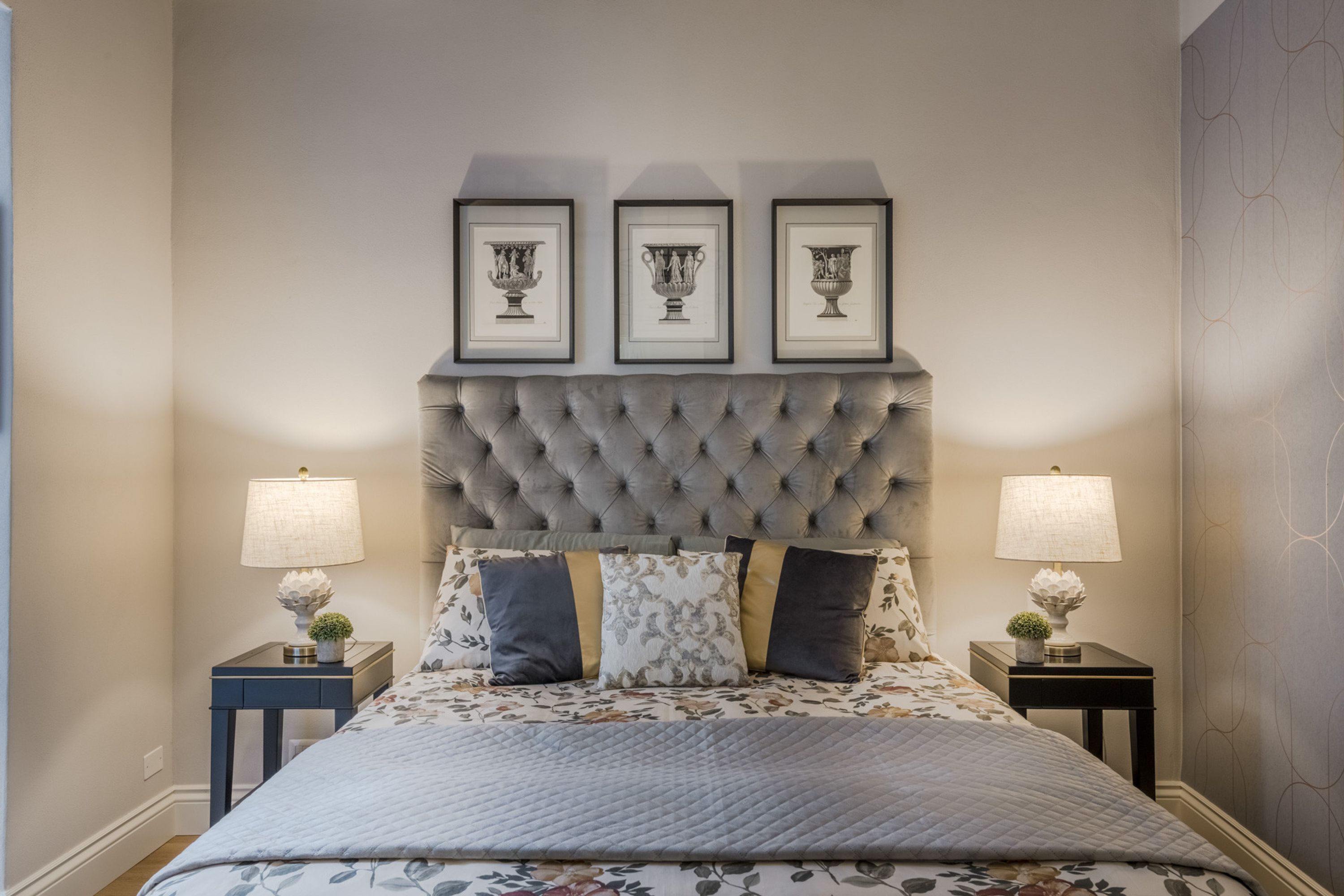 A bedroom with a tufted headboard, a floral bedspread, and three framed pictures above the bed. There are two bedside tables with lamps and decorative items.