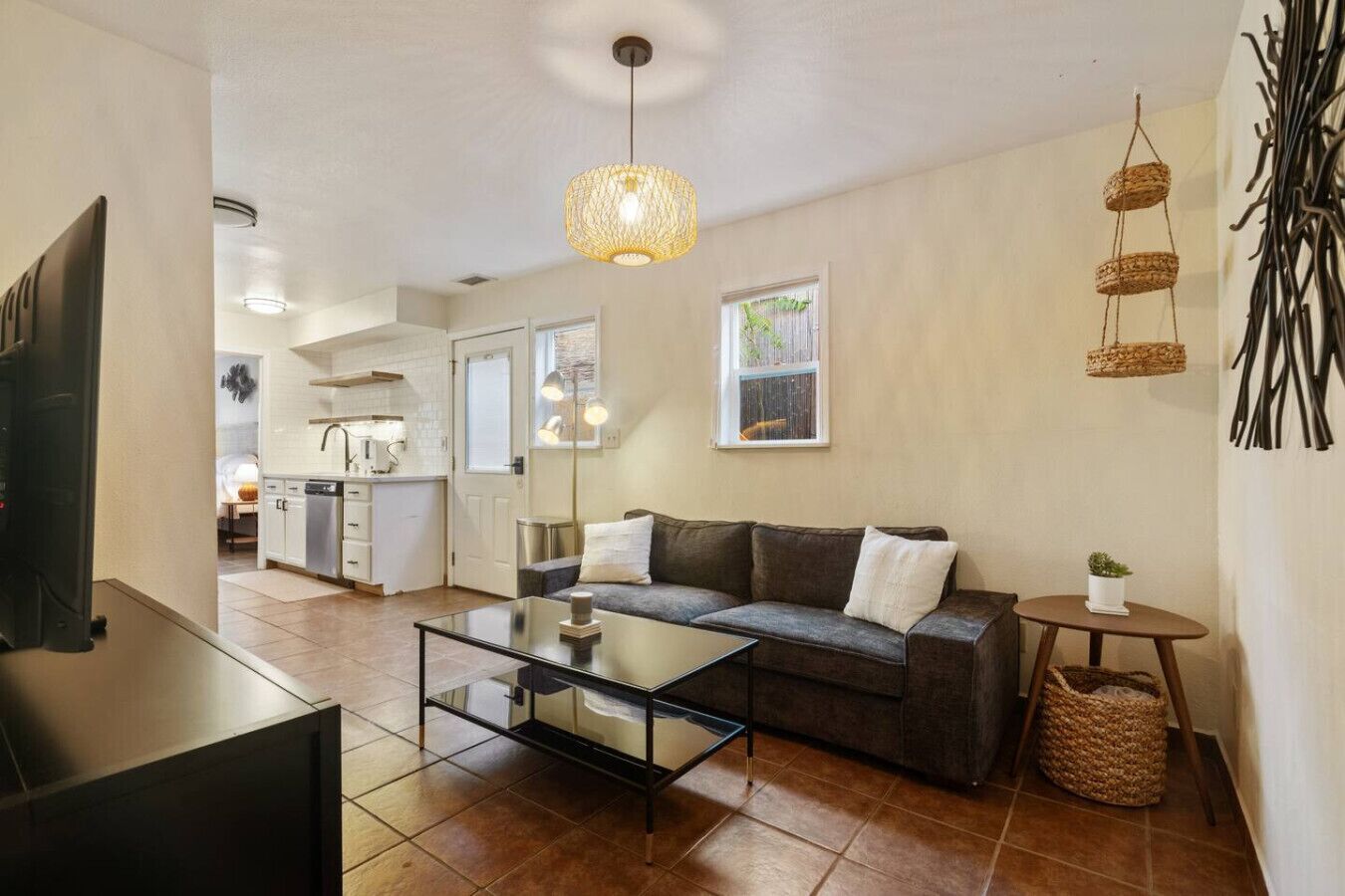 Een moderne woonkamer met een bank, salontafel en televisie. In de achtergrond is een keuken met witte kastjes en een raam.