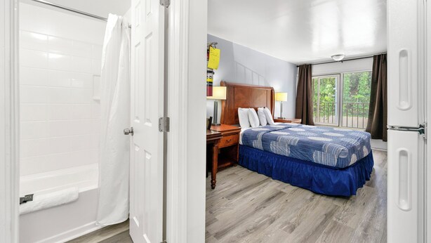 Ein Hotelzimmer mit Holzboden, einem Bett mit blauer Bettdecke, einem Nachttisch, einem Schreibtisch, einer Lampe und einem Fenster mit Vorhängen.