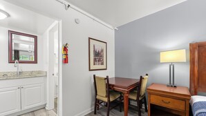 Ein Zimmer mit Holztisch, Stühlen, einem kleinen Nachttisch mit Lampe, einem gerahmten Bild an der Wand und einem Feuerlöscher.