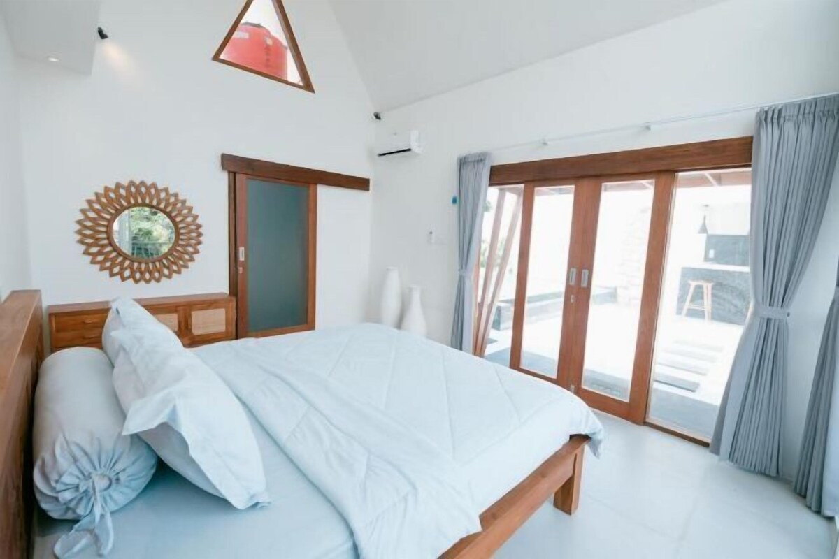 Une chambre à coucher avec un lit, une tête de lit en bois, une grande fenêtre avec des rideaux et un objet décoratif fixé au mur.