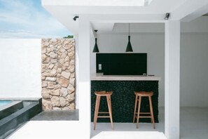 A modern outdoor kitchen with a stone wall, two bar stools, and a TV.