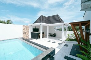 A modern outdoor pool area with a covered seating area, stone wall, and a clear blue sky.