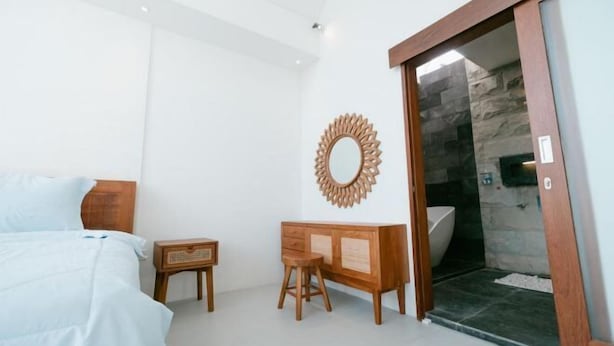 A bedroom with a bed, wooden nightstand, dresser, and a bathroom with a bathtub and stone wall. - The Lavana Hidden Villa Lombok (Mataram)