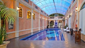 An indoor swimming pool with a glass ceiling, surrounded by arched windows and potted plants.