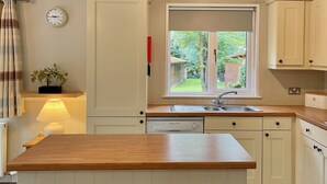 A kitchen with a wooden island, two stools, a clock, a window with a view of greenery, and a ceiling light fixture. - Holiday Lodge in The Cotswolds (Kingham)