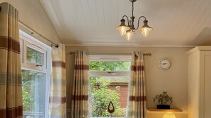 A dining area with a wooden table, white chairs, a chandelier, a window with curtains, and a clock on the wall. - Holiday Lodge in The Cotswolds (Kingham)