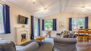 A living room with a fireplace, grey sofas, a wooden coffee table, and a dining area with a table and chairs. - Holiday Lodge in The Cotswolds (Kingham)