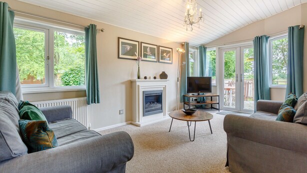 A living room with two sofas, a fireplace, a TV, and large windows with curtains. - Holiday Lodge in The Cotswolds (Kingham)