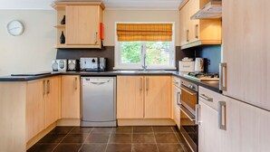 A modern kitchen with wooden cabinets, a dishwasher, a stove, and a window with a view of greenery. - Holiday Lodge in The Cotswolds (Kingham)
