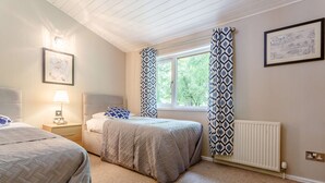 A bedroom with two beds, a window with patterned curtains, a radiator, and a wall-mounted lamp. - Holiday Lodge in The Cotswolds (Kingham)