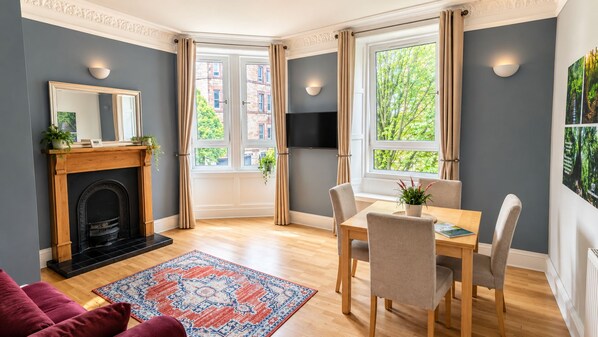A room with a fireplace, a dining table with chairs, a patterned rug, and a large window with curtains.