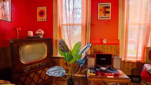 A room with a red wall, a vintage radio, a plant, a small table with a laptop, and a window with curtains. - 4 BR Home 5 Mins to Cedar Point, Retro 1970s! (Sandusky)