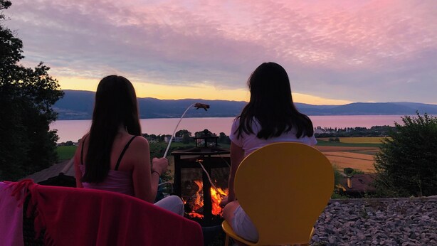 Two people sitting by a fire, overlooking a lake and mountains.