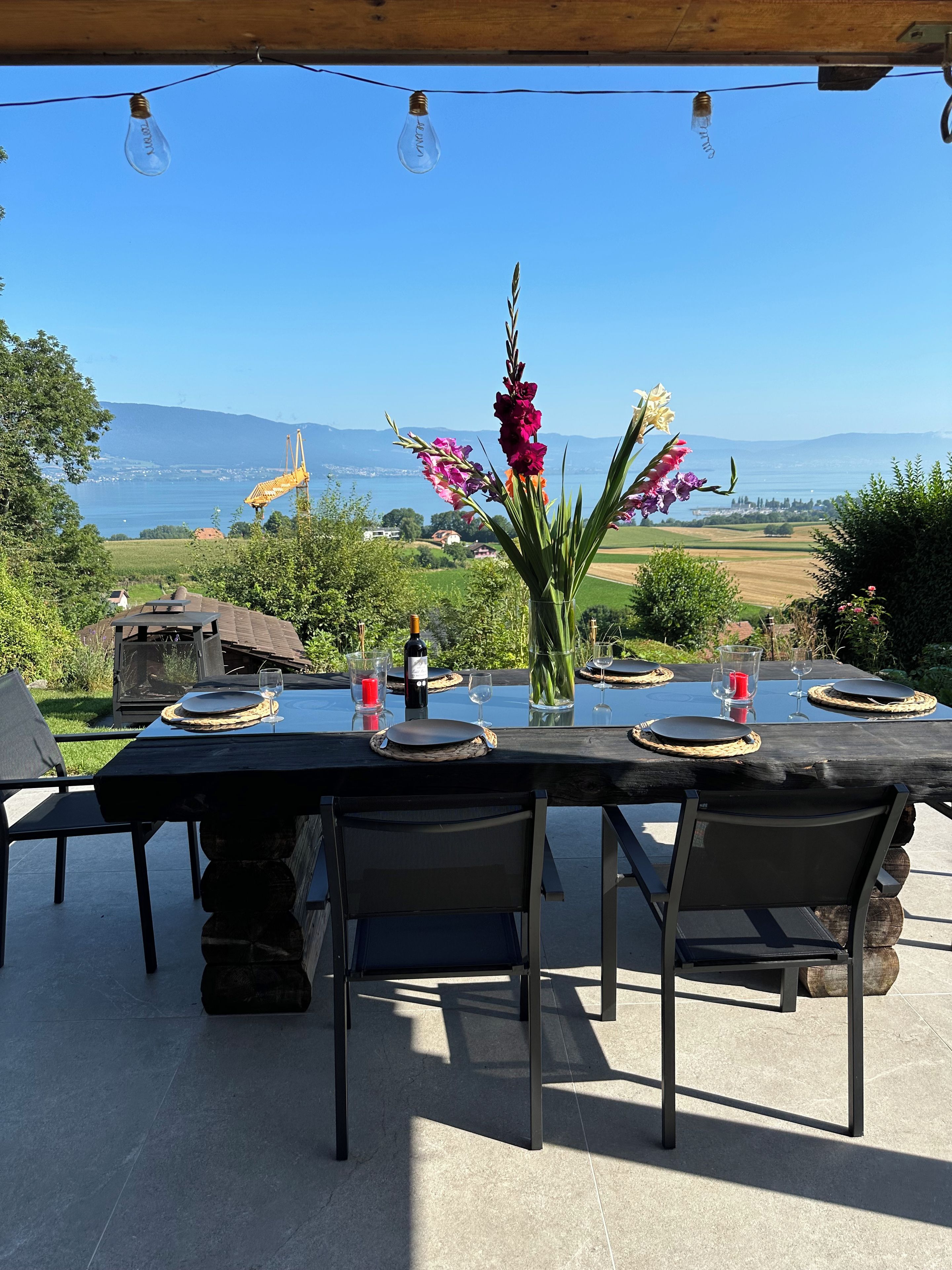 Un espace de restauration en plein air avec une table dressée pour quatre personnes, offrant une vue panoramique sur un paysage magnifique, avec des montagnes et une étendue d’eau.