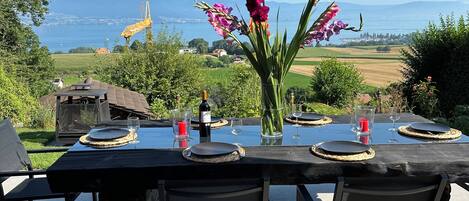 Un espace de restauration en plein air avec une table dressée pour quatre personnes, offrant une vue panoramique sur un paysage magnifique, avec des montagnes et une étendue d’eau.