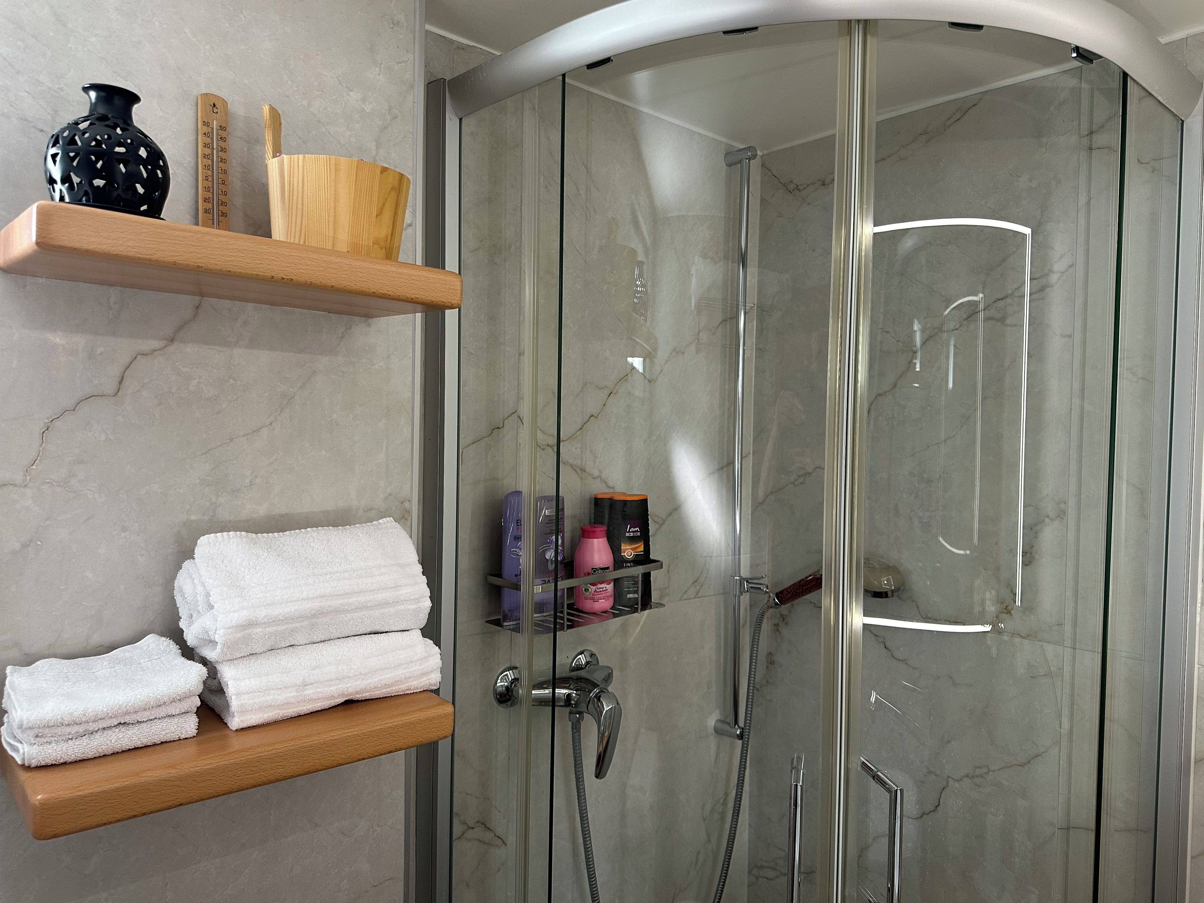 A modern bathroom with a glass-enclosed shower, marble walls, and wooden shelves holding towels and toiletries.