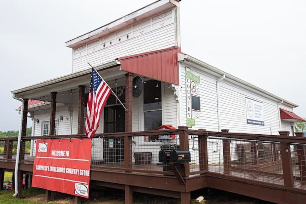 A white building with a porch, an American flag, and a red banner reading "Welcome to Campbell's Creek Country Store."