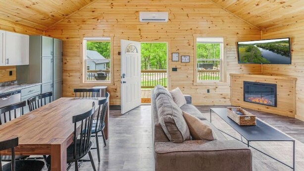 A wooden cabin interior with a dining area, sofa, fireplace, and a TV mounted on the wall. - Hot Tub~Firepit~Riverfront~Near Lake Cumberland (Jamestown)