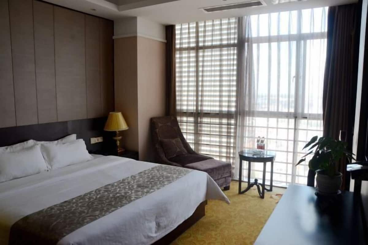 A hotel room with a large bed, a chair, a small table, and a window with blinds.