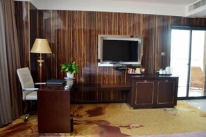 Une chambre d’hôtel avec un mur recouvert de panneaux de bois, un téléviseur à écran plat fixé au-dessus d’un bureau en bois foncé, une chaise blanche, une plante en pot et une petite table avec une lampe.