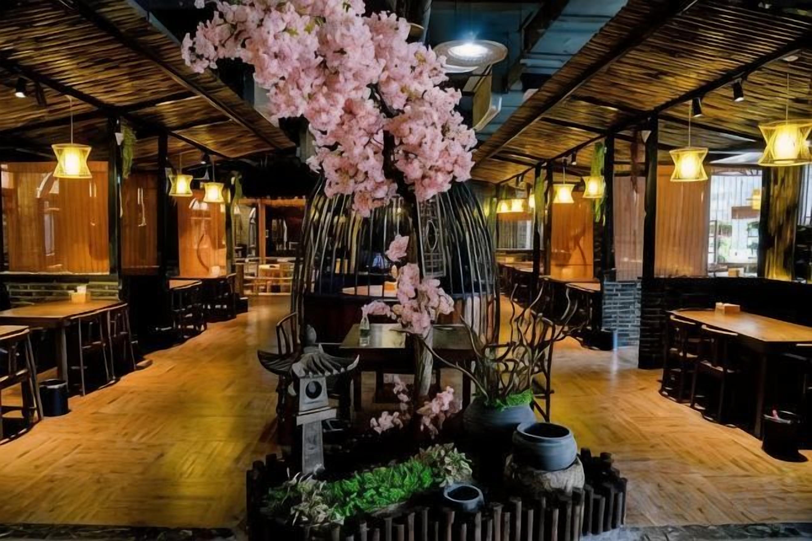 A restaurant interior with wooden flooring, tables, chairs, hanging lights, and a central decorative tree.