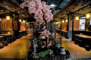 Interior de un restaurante con suelo de madera, mesas, sillas, luces colgantes y un árbol decorativo en el centro.