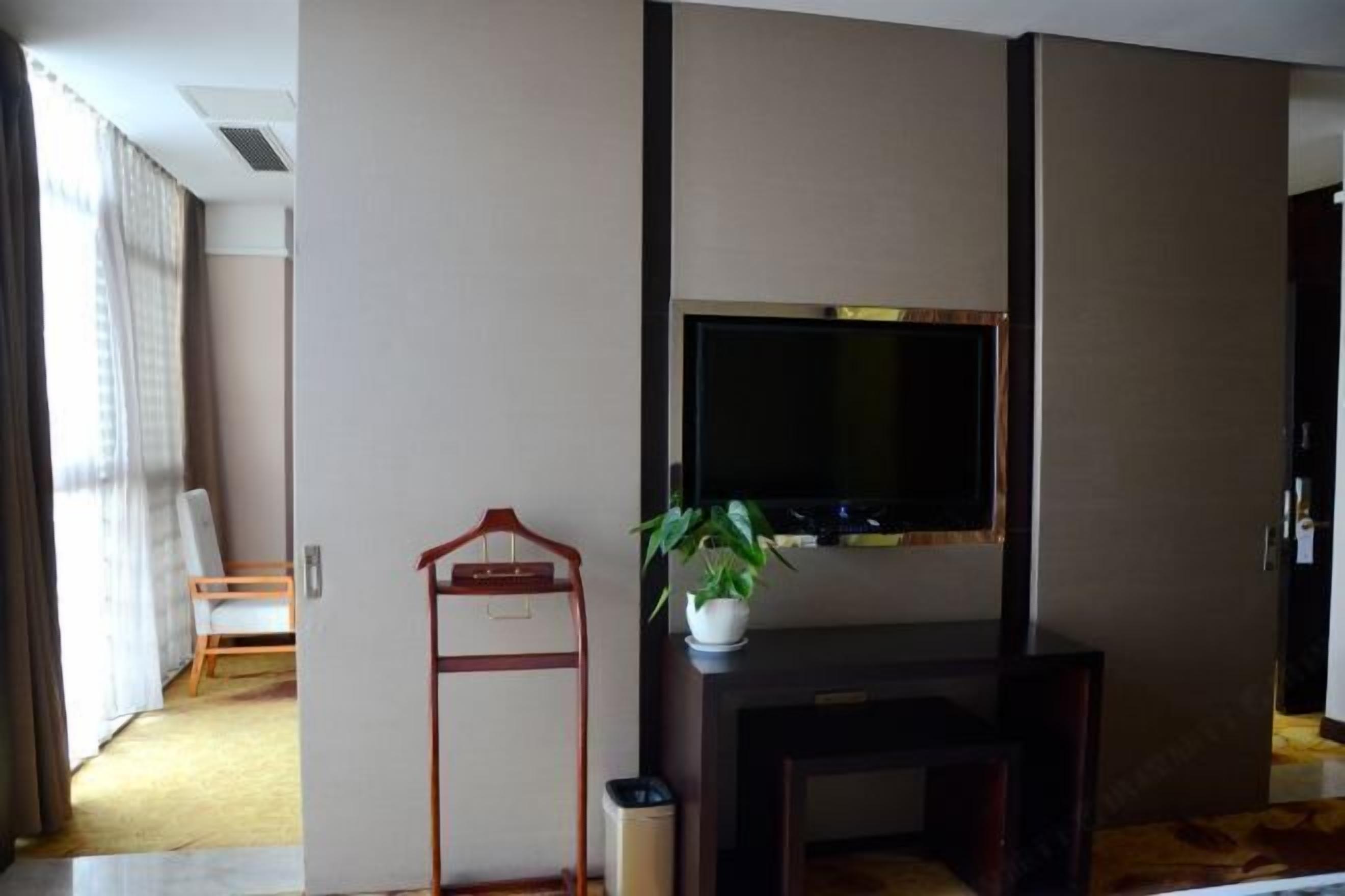 Habitació d'hotel amb televisió de pantalla plana penjada a una càmera de fusta fosca, una planta en test, una penjador de fusta i una cadira blanca al fons.