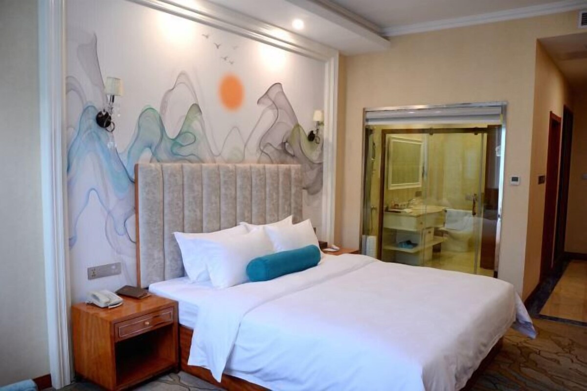 A hotel room with a bed, a nightstand, a wall with abstract art, a bathroom with a glass door, and a hallway.