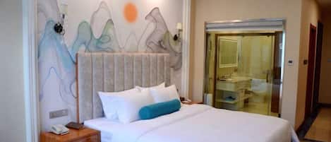 A hotel room with a bed, a nightstand, a wall with abstract art, a bathroom with a glass door, and a hallway.