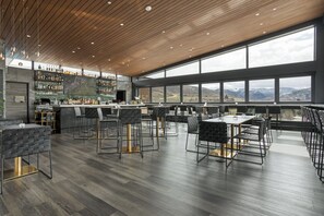 A modern bar area with a wooden ceiling, large windows offering a mountain view, and a variety of bar stools and tables.