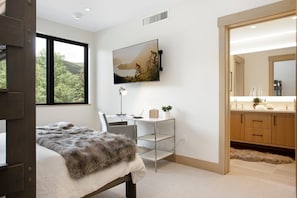 Un dormitorio con cama, escritorio, silla, televisor montado en la pared y baño con espejo y lavamanos.