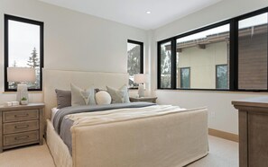 A modern bedroom with a large bed, bedside tables, and a dresser. There are two windows providing a view of the outside.