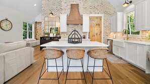A kitchen with a large island, white cabinetry, and a brick wall. There are three wicker bar stools around the island. - Hickory Cottage - 1bdrm Retreat (Taylors)
